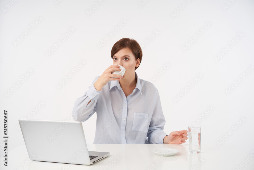 Indoor shot of young brown-eyed short haired brunette female with natural makeup drinking coffee while working in modern office with her laptop, isolated over white background Indoor shot of young brown-eyed short haired brunette female with natural makeup drinking coffee while working in modern office with her laptop, isolated over white background