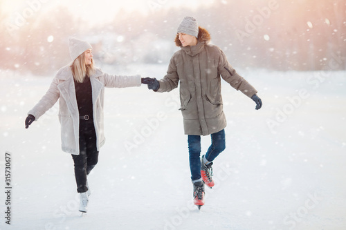 Loving couple in winter with skates on rink on sunset background, guy and girl dressed in warm hat