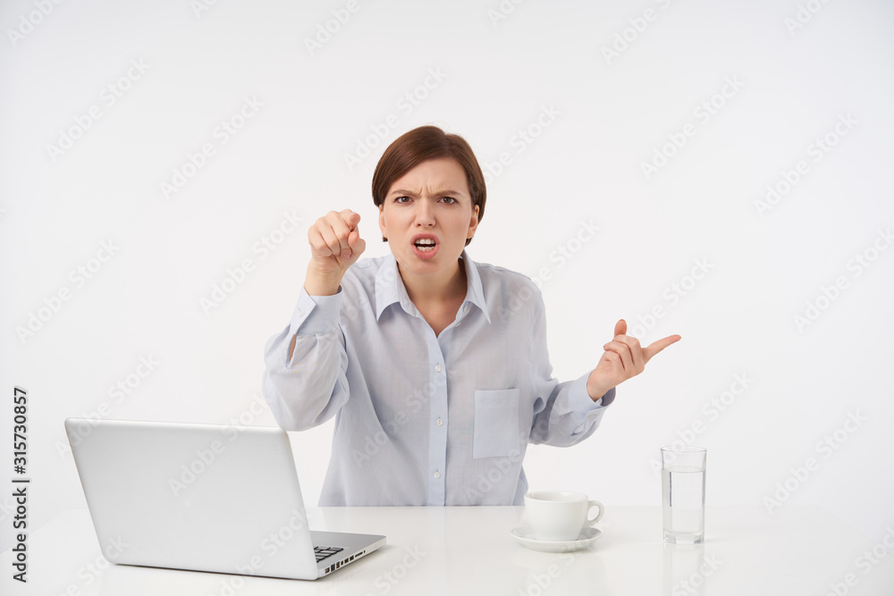 Disgruntled young brown-eyed short haired brunette lady with casual hairstyle showing with forefinger at camera and frowning angrily her face, isolated over white background Disgruntled young brown-eyed short haired brunette lady with casual hairstyle showing with forefinger at camera and frowning angrily her face, isolated over white background