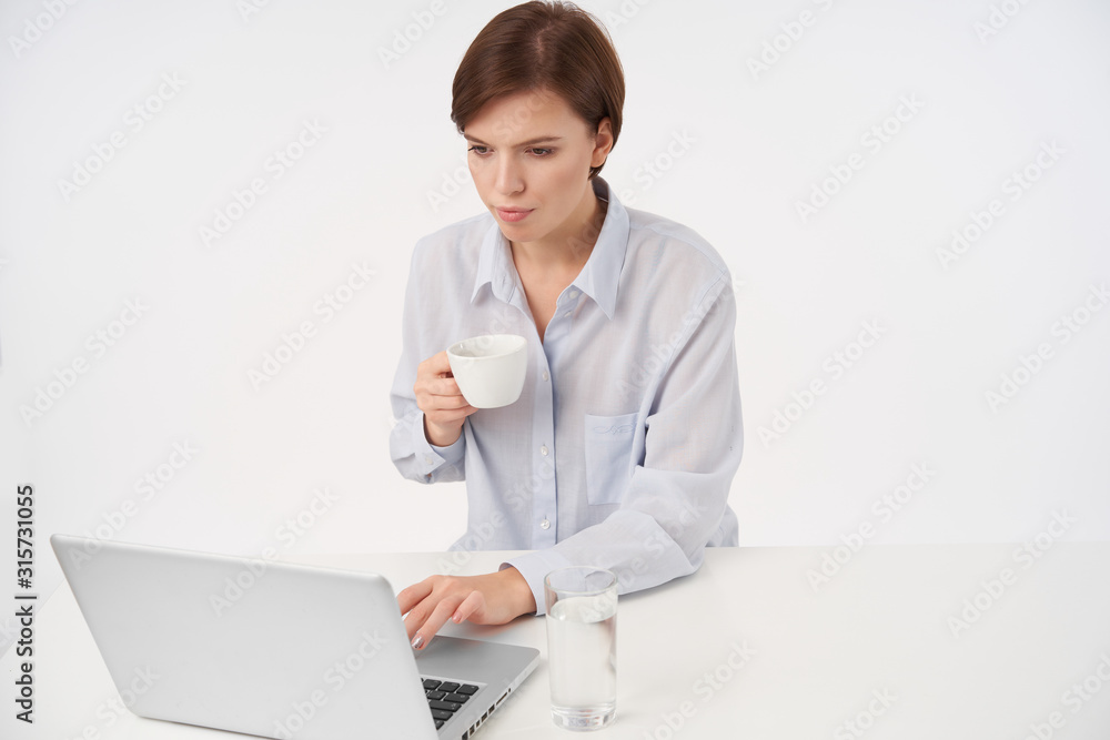 Indoor photo of young pretty short haired brunette woman with casual hairstyle keeping hand on keyboard of laptop and looking at screen with concentrated face, isolated over white background Indoor photo of young pretty short haired brunette woman with casual hairstyle keeping hand on keyboard of laptop and looking at screen with concentrated face, isolated over white background