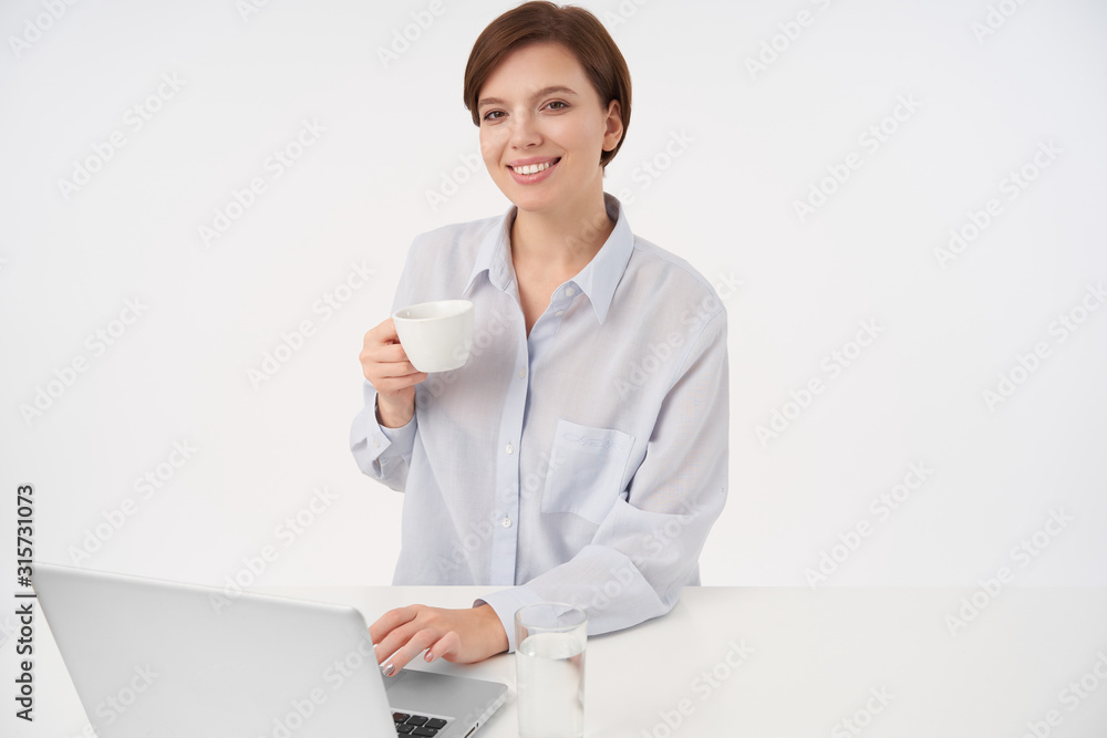 Charming young short haired brunette female lady with cup of coffee in raised hand looking cheerfully at camera and smiling pleasantly, dressed in formal clothes over white background Charming young short haired brunette female lady with cup of coffee in raised hand looking cheerfully at camera and smiling pleasantly, dressed in formal clothes over white background