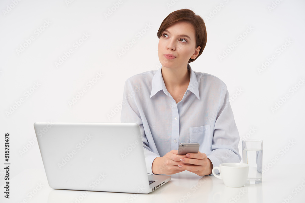 Pensive young pretty short haired brunette lady with short trendy haircut looking thoughtfully aside with folded lips and keeping smartphone in hands, isolated over white background Pensive young pretty short haired brunette lady with short trendy haircut looking thoughtfully aside with folded lips and keeping smartphone in hands, isolated over white background