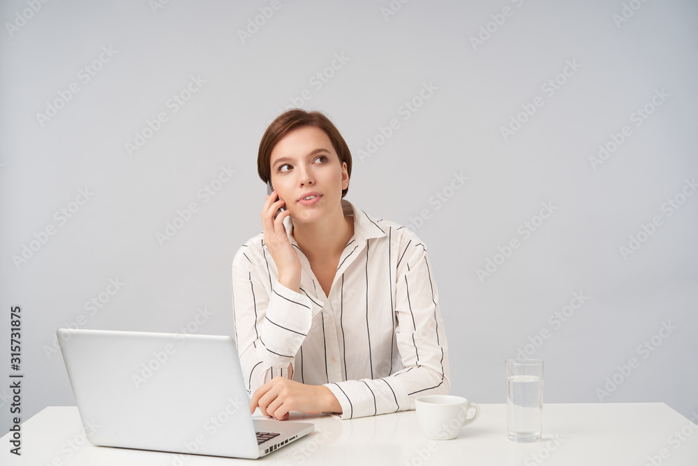 Indoor shot of busy young pretty brown haired lady with natural makeup working in modern office with her laptop, holding mobile phone and making call while posing over white background Indoor shot of busy young pretty brown haired lady with natural makeup working in modern office with her laptop, holding mobile phone and making call while posing over white background