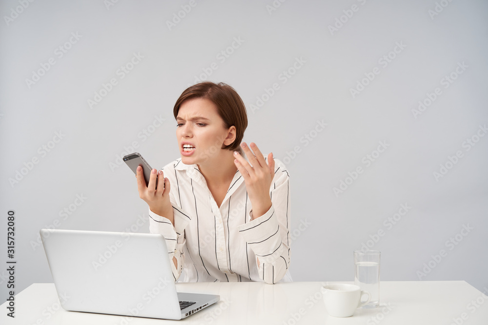 Stressed young brown haired pretty business woman having hard day at her work, holding mobile phone in raised hand and looking angrily on it, isolated over white background Stressed young brown haired pretty business woman having hard day at her work, holding mobile phone in raised hand and looking angrily on it, isolated over white background