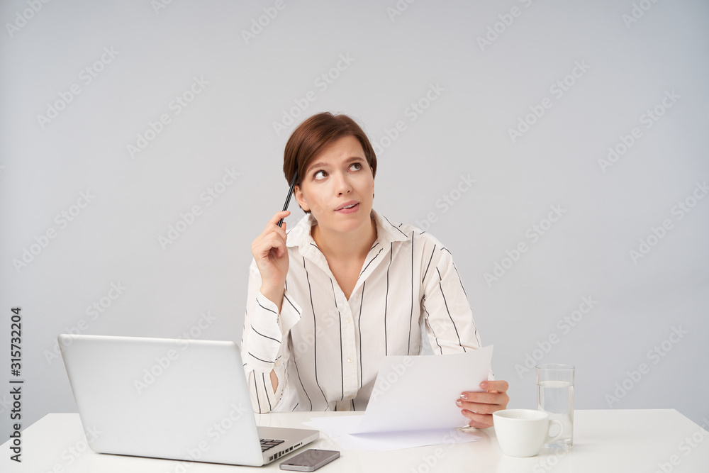 Indoor photo of young short haired brunette female employee holding piece of paper and looking thoughtfully upwards, preparing documents for upcoming meeting, isolated over white background Indoor photo of young short haired brunette female employee holding piece of paper and looking thoughtfully upwards, preparing documents for upcoming meeting, isolated over white background