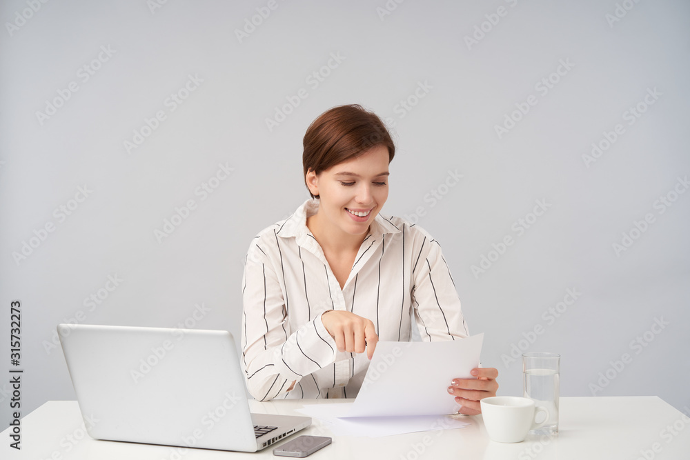 Cheerful young pretty brown haired lady in formal clothes sitting at table over white background, reading text on piece of paper and being pleased with it Cheerful young pretty brown haired lady in formal clothes sitting at table over white background, reading text on piece of paper and being pleased with it