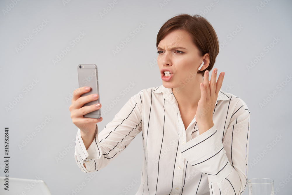Displeased young pretty short haired brunette female frowning her eyebrows and looking at screen with pout while having stressful phone conversation, posing over white background Displeased young pretty short haired brunette female frowning her eyebrows and looking at screen with pout while having stressful phone conversation, posing over white background