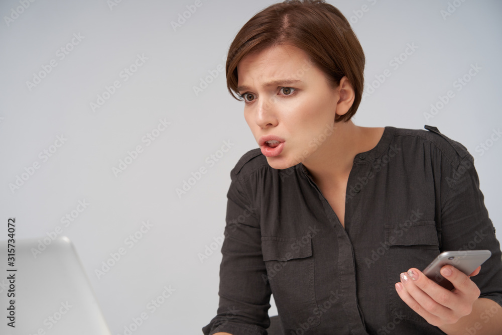 Close-up of young confused brown-eyed pretty brunette lady with short trendy haircut keeping smartphone in raised hand and frowning eyebrows while looking in front of herself Close-up of young confused brown-eyed pretty brunette lady with short trendy haircut keeping smartphone in raised hand and frowning eyebrows while looking in front of herself