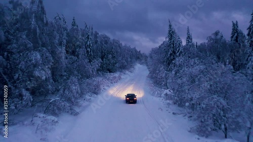 Aerial: flying above car driving alone through a snowy frozen forest at cloudy dark evening. Empty rural road surrounded by snow covered trees. 