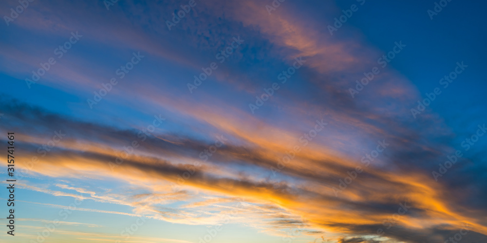 Panoramic orange illuminated sky background in evening dusk view. Dramatic sunset on blue striped cloudscape with sunlit fiery clouds. Scenic natural panorama of afterglow light. Ecology and weather.