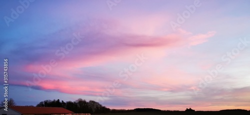 A fantastic sunset with red yellow orange and purple tones with dramatic cloud formation over green meadows with bare trees in January on the island of Rügen