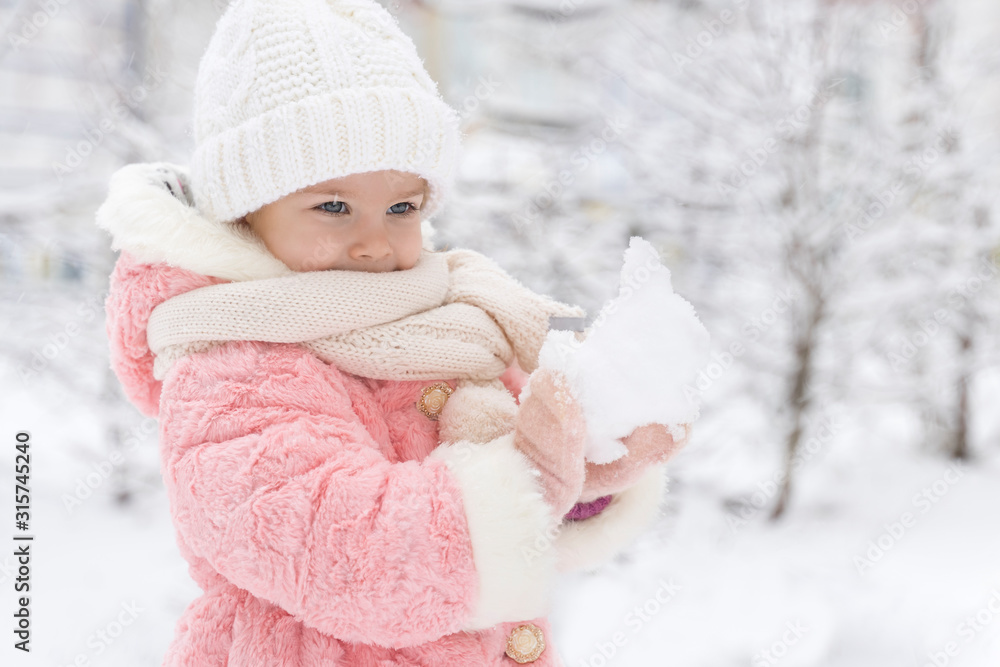 beautiful girl in a pink fur coat smiles and sculpts a snowball in a snowy yard