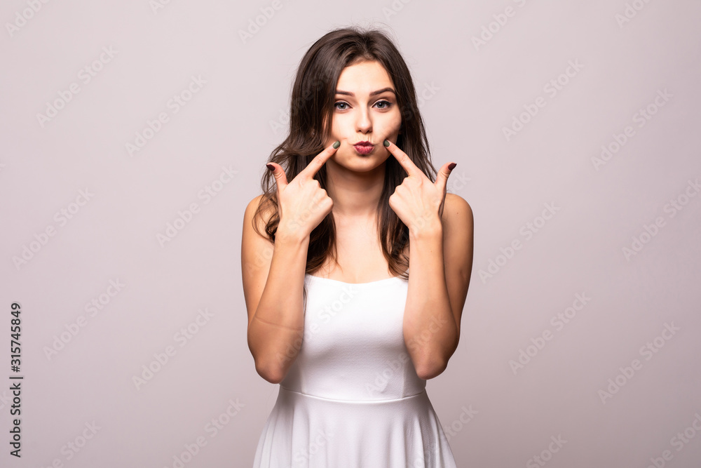 Amusing woman pointing fingers at cheeks and grimacing isolated over white background Amusing woman pointing fingers at cheeks and grimacing isolated over white background