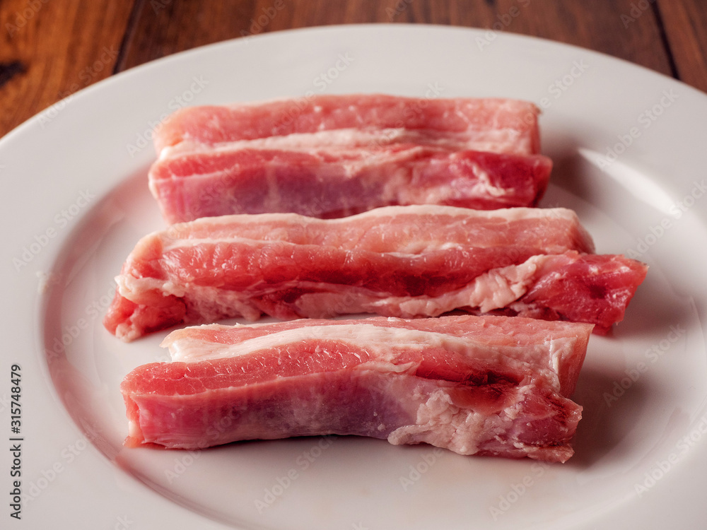 Uncooked raw pork belly slices on a white plate and wooden table. Uncooked raw pork belly slices on a white plate and wooden table.