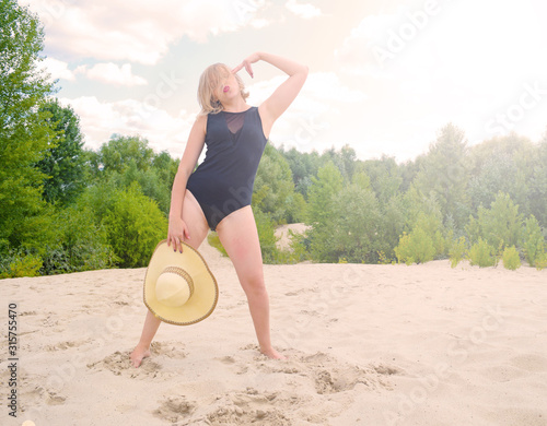 A young woman in a black bathing suit with a hat stands on the sand in the desert.