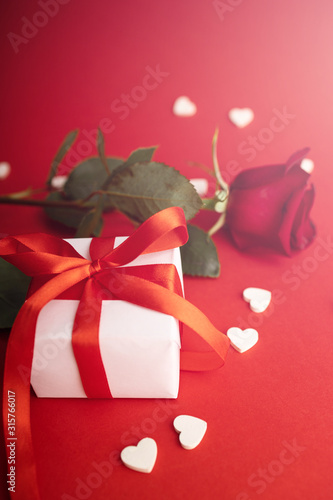 Fototapeta Naklejka Na Ścianę i Meble -  Red rose Gift for valentines day. View of present box decorated into the white paper with red bow-knot with signs of white wooden hearts besides. Flat lay on red background. Love and amour.
