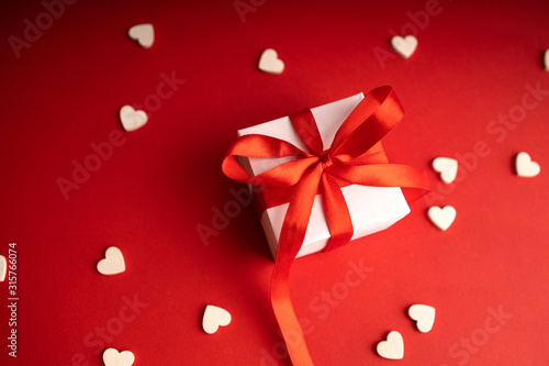 Top view of present box package decorated into the white paper and red bow-knot with signs of white wooden hearts besides. Flat lay on red background. Concept of celebrate.