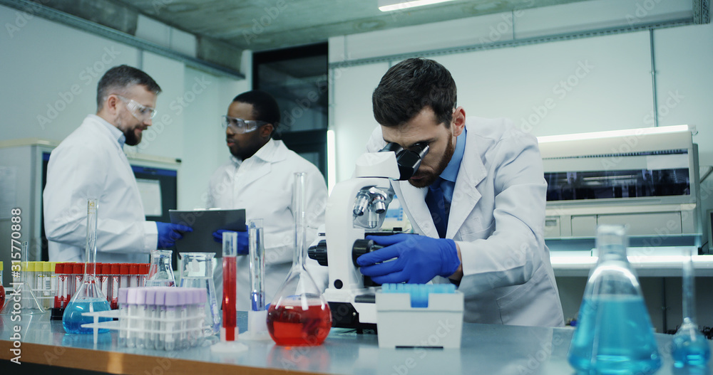 Portrait shot of the Caucasian man scientist looking in the microscope while investigating something in the laboratory, while his male mixed-races co-workers talking on the background. Portrait shot of the Caucasian man scientist looking in the microscope while investigating something in the laboratory, while his male mixed-races co-workers talking on the background.