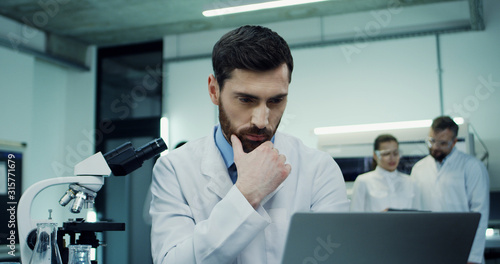 Portrait of the handsome Caucasian man scientist doing some investigation at the laptop computer and analysis while looking in the microscope in the laboratory.