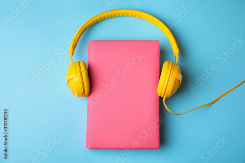 Book with blank cover and headphones on light blue background, flat lay