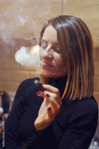 Принти на полотні Mature caucasian woman at kitchen home smoking tobacco holding a cigarillo weari