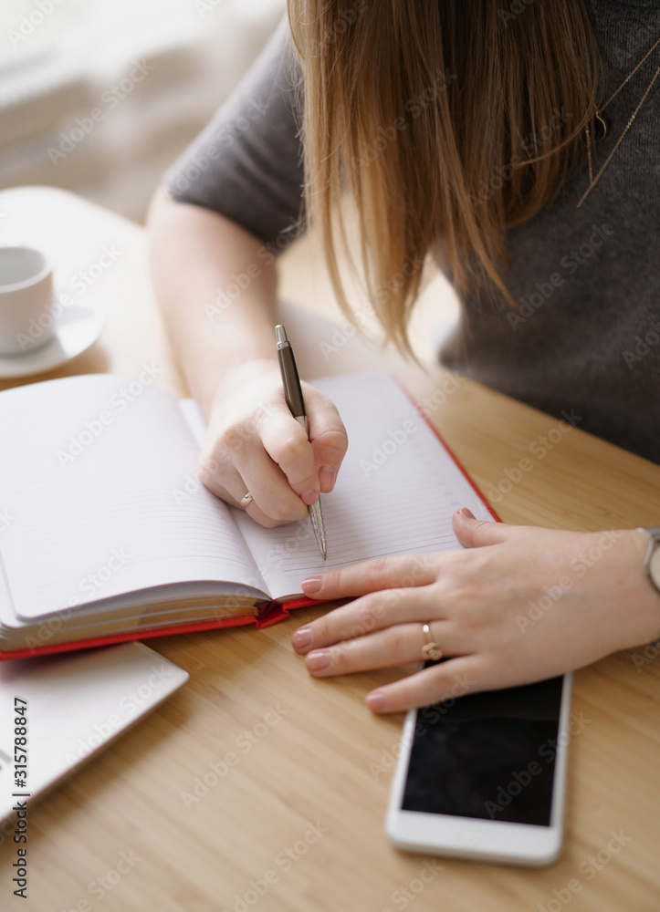 Woman writes in a red notebook sitting at a wooden table with a cup of coffee. Nearby lies a smartphone. Communication in business. Woman writes in a red notebook sitting at a wooden table with a cup of coffee. Nearby lies a smartphone. Communication in business.