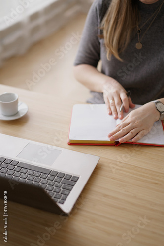 Woman writes in a red notebook sitting at a wooden table with a cup of coffee. Nearby lies a laptop. Communication in business.