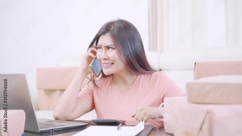 Online shop owner making a phone call and looks angry while working with laptop and cardboard on the table in the living room at home. Shot in 4k resolution Online shop owner making a phone call and looks angry while working with laptop and cardboard on the table in the living room at home. Shot in 4k resolution