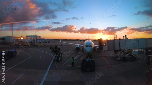 Airport Sunset Time-lapse. A plane is moved from the gate toward the runway. Workers shuffle around below, preparing the aircraft. The sun goes down and the clouds burn orange. 4K.