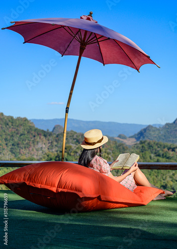 young woman sitting on pillow sofa outdoor on balcony terrace of the cefe, enjoy mountain view with offline social media by reading a book comfortable