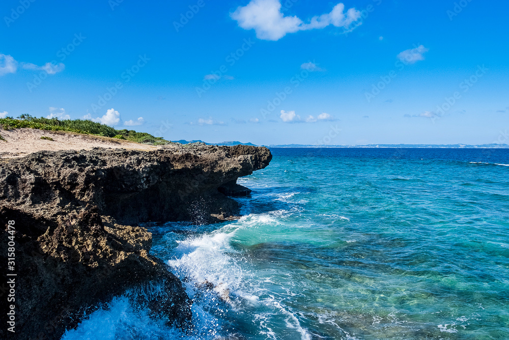 青く透き通った海の海岸沿いの岩場に波しぶきが上がっている晴れた日の久高島の風景