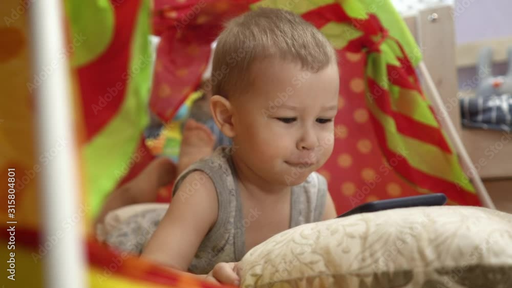 Charming baby plays with the phone in the children's tent. He watches videos and pictures. Charming baby plays with the phone in the children's tent. He watches videos and pictures.
