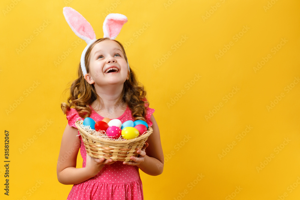 Beautiful cute little girl in Easter bunny ears holds a basket with eggs on a yellow background. The child raised his head and looks up. Copy space
