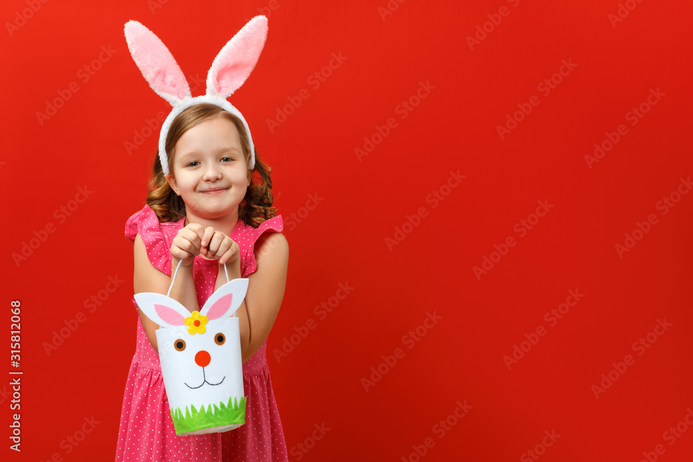 Beautiful cute little girl in Easter bunny ears holds a bag for sweets and eggs. Portrait of a child on a red background. Copy space Beautiful cute little girl in Easter bunny ears holds a bag for sweets and eggs. Portrait of a child on a red background. Copy space