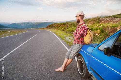 Portrait of a bearded happy serious traveler hipster with a backpack in a plaid shirt and a hat next to an unknown car stands on the road at sunset in the mountains. Happy and confident travel concept