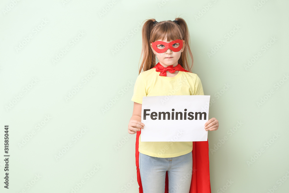 Little girl in superhero costume holding paper with word FEMINISM on color background