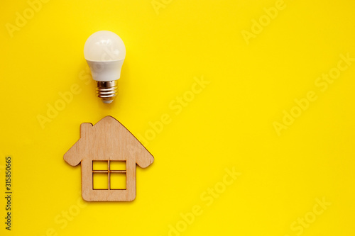 LED light bulb and wooden house on yellow background with copy space. Top view, flat lay. Energy save concept