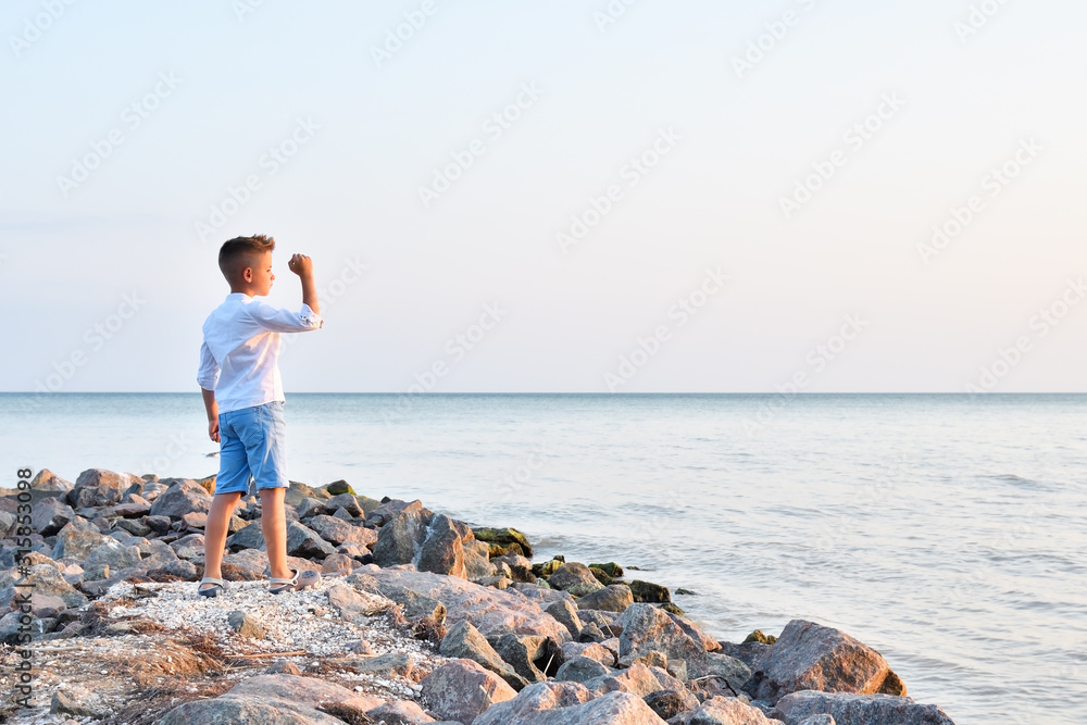 Beautiful boy standing on the stones on the seashore watching the sunset