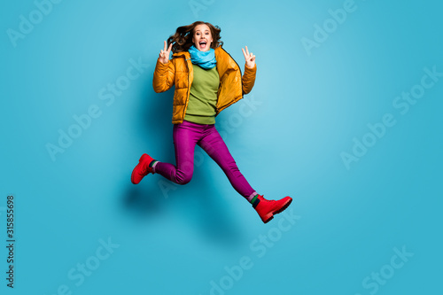 Full length photo of pretty lady jump high up showing v-sign symbols wear casual yellow overcoat scarf violet pants red boots green pullover isolated blue color background