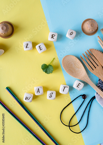 Zero Waste concept. Wooden spoon, fork, knife, bamboo cocktail tube and metal tubules on a blue and yellow background with wooden cubes with the inscription zero waste