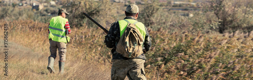 A mans with a gun in his hands and an camouflage vest on a pheasant hunt in a wooded area in cloudy weather. Hunters in search of game.
