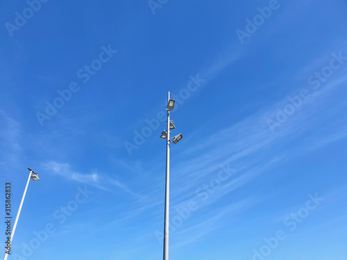 Tall metal poles with street lights are seen from below. One pole has four lights, the other one. The sky is blue with trails of faint white clouds.