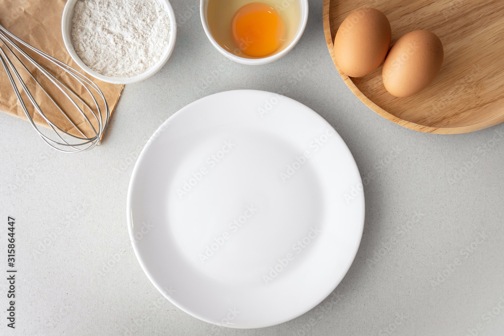 Top view above getting ready to bake, whisk, bowl of flour, small bowl of cracked egg white egg yolk, and raw eggs with empty white plate on grey background. Top view above getting ready to bake, whisk, bowl of flour, small bowl of cracked egg white egg yolk, and raw eggs with empty white plate on grey background.