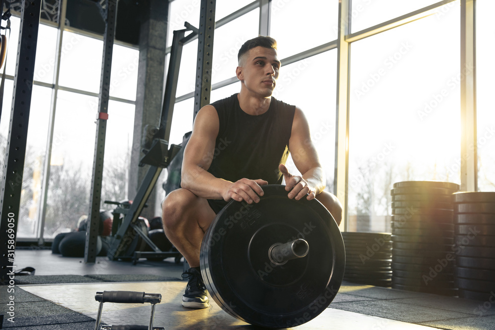 Confident. Young muscular caucasian athlete training in gym, doing strength exercises, practicing, work on his upper body with weights and barbell. Fitness, wellness, healthy lifestyle concept. - obrazy, fototapety, plakaty 