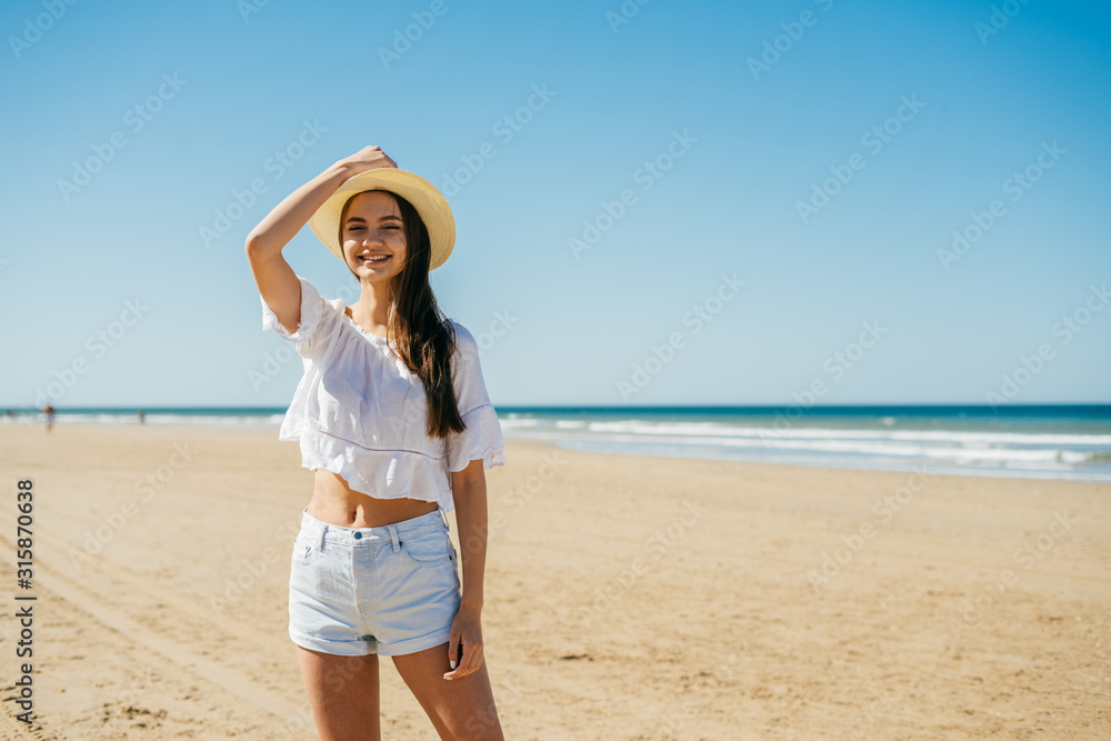 girl in a hat and light clothes posing on a deserted beach on the background of the ocean
