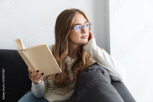 Photo of young serious woman reading book while sitting on couch