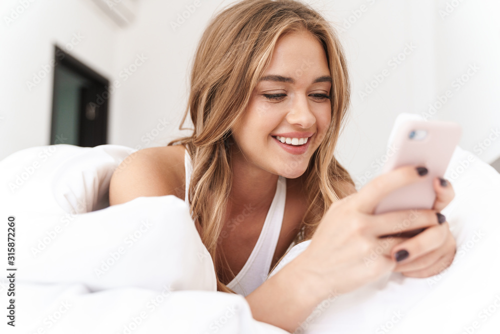 Photo of smiling caucasian woman using cellphone while lying in bed Photo of smiling caucasian woman using cellphone while lying in bed