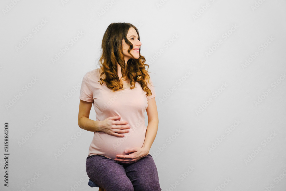 Attractive pregnant young woman sitting against grey wall. Happy pregnancy concept. - obrazy, fototapety, plakaty 