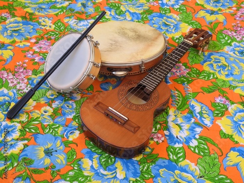 Three Brazilian musical instruments: cavaquinho, pandeiro (tambourine) and tamborim with drumstick on a colorful “chitao” fabric with floral prints. They are widely used in samba and pagode ensembles.