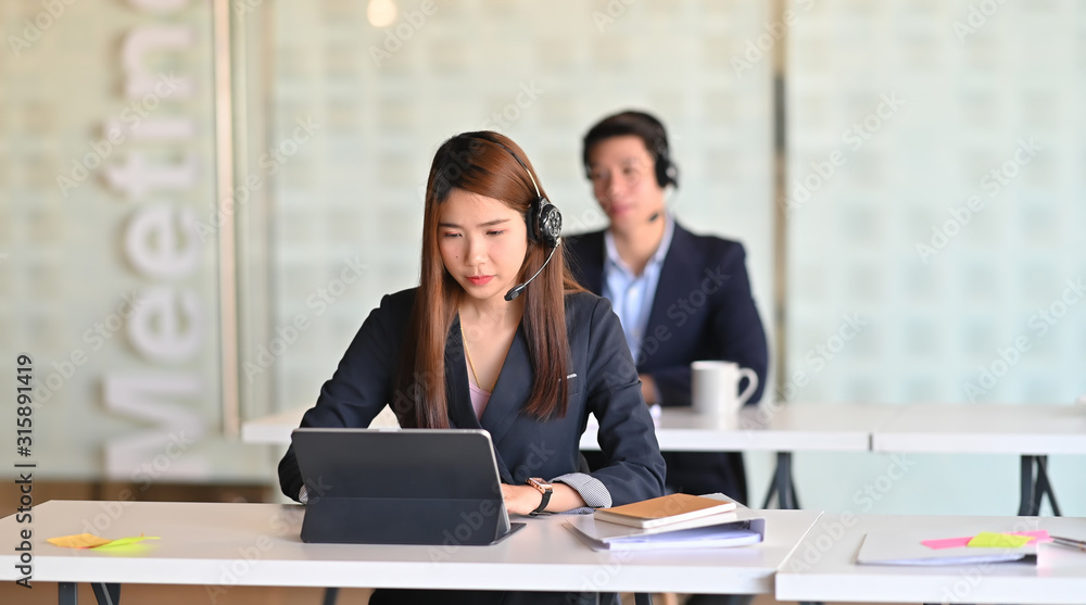 Beautiful call center woman in black suit she looking on tablet screen and sitting at work desk in front of her colleague in the modern office. Beautiful call center woman in black suit she looking on tablet screen and sitting at work desk in front of her colleague in the modern office.